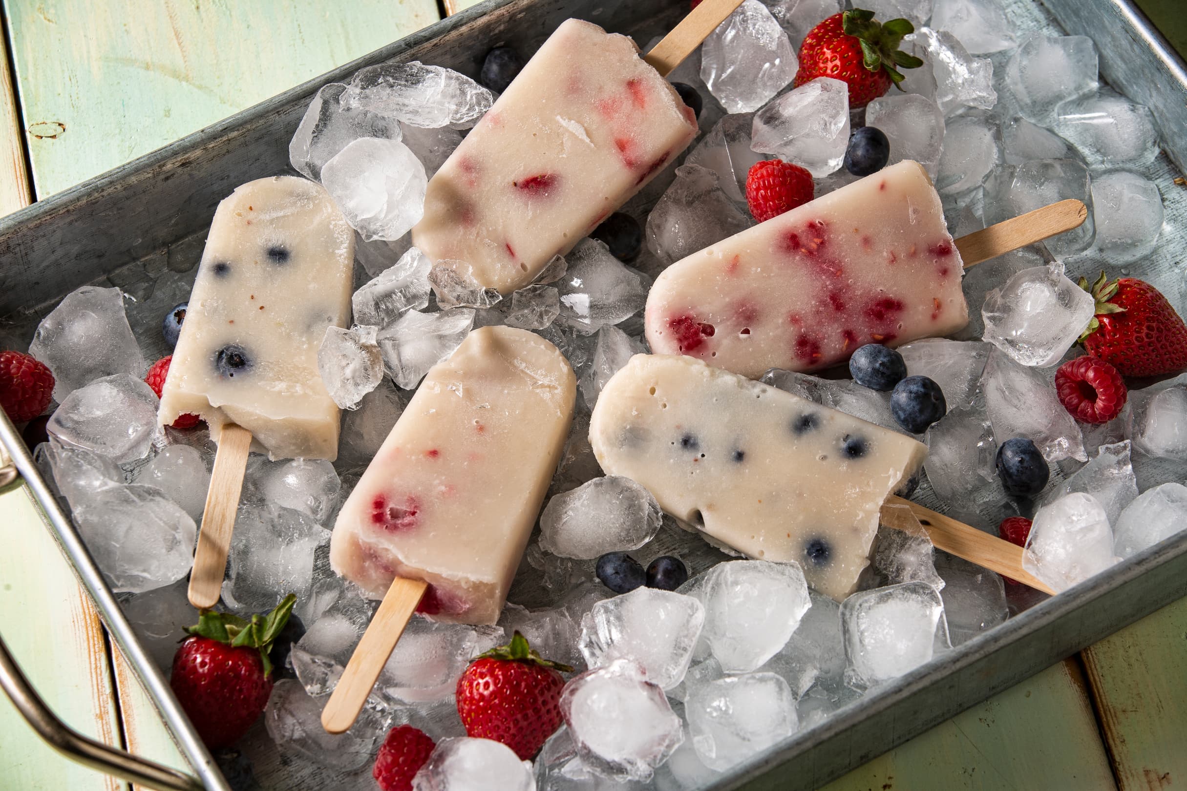 Five berry-filled popsicles are arranged on ice in a metal tray, surrounded by fresh strawberries and blueberries.
