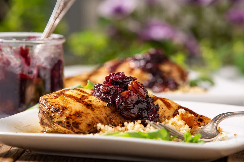Grilled chicken breast topped with a dark berry sauce, served on a bed of couscous with greens on a white plate; a jar of sauce is in the background.
