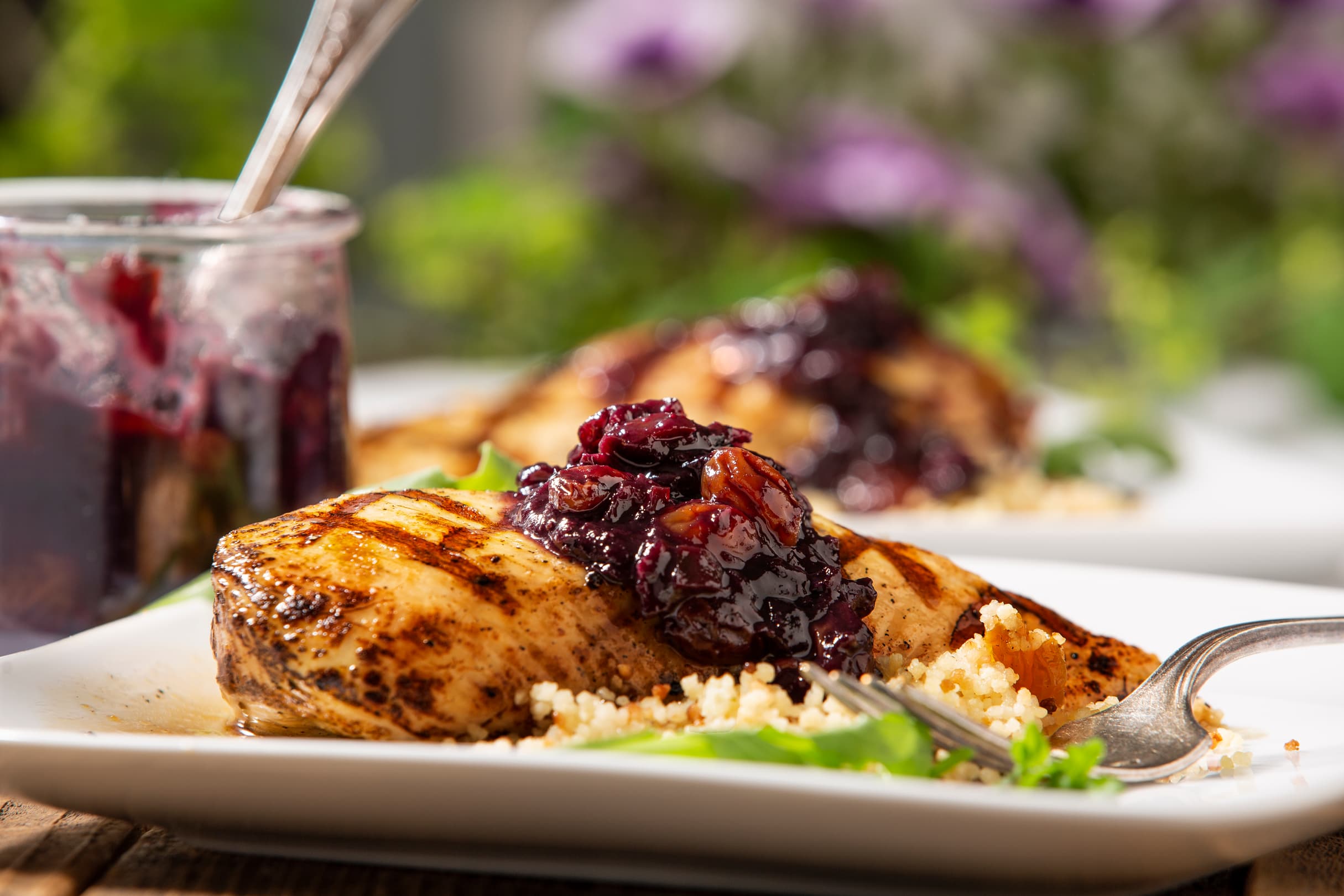 Grilled chicken breast topped with a dark berry sauce, served on a bed of couscous with greens on a white plate; a jar of sauce is in the background.