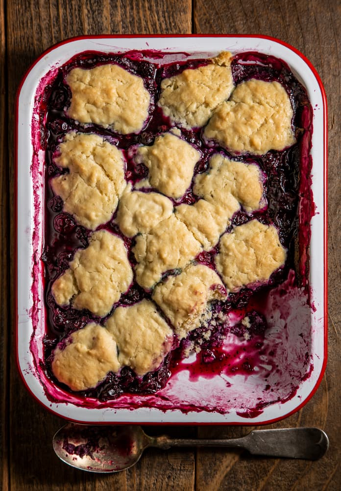 A baked berry cobbler in a white and red baking dish, topped with golden biscuit dough, sits on a wooden table next to a used spoon.