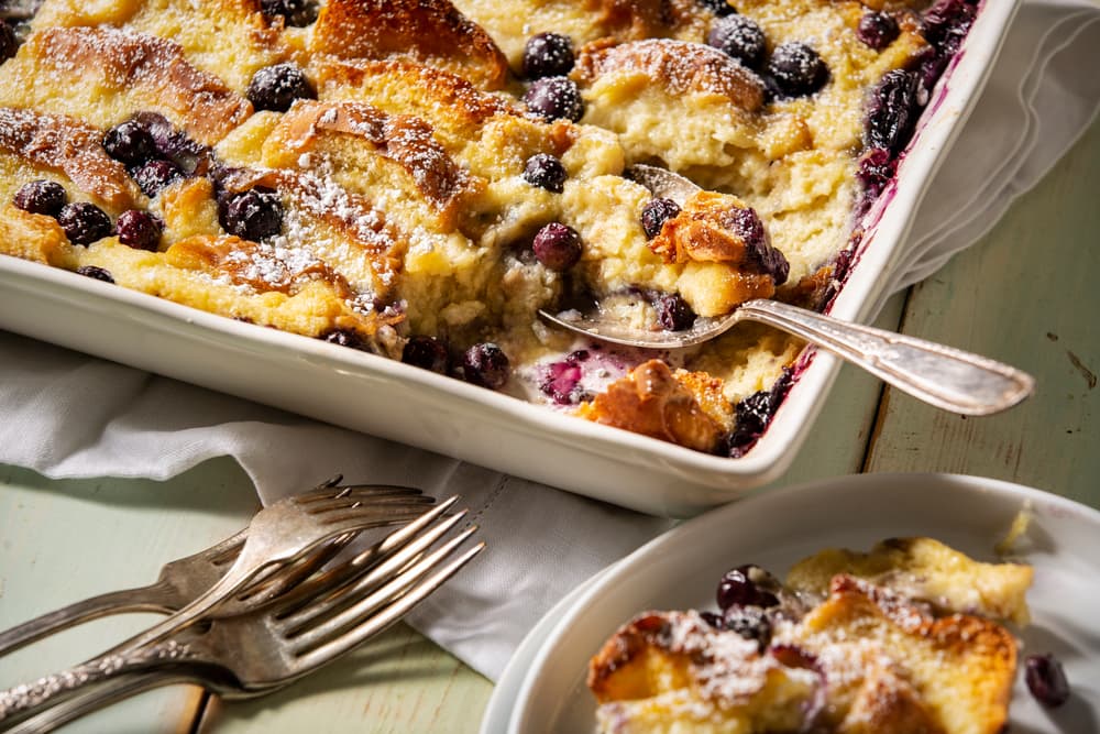 A baking dish of blueberry bread pudding with a serving spoon, some pieces missing, and a plate with a portion. Four forks are placed nearby on the table.