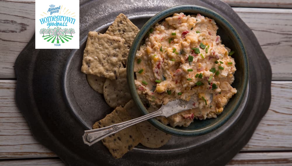 A bowl of pimento cheese spread with chopped chives, served with crackers on a dark plate. A Hometown Goodness Tour logo appears in the top left corner.