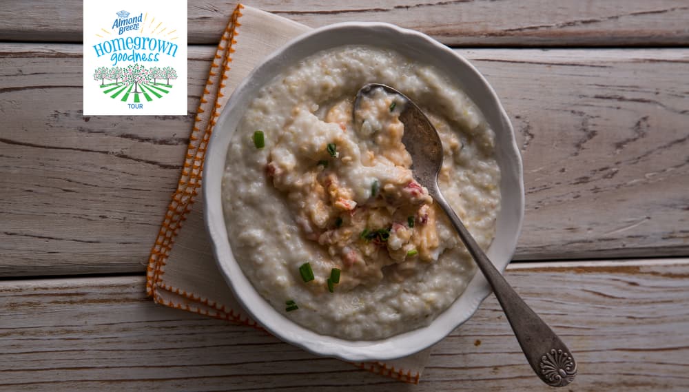 A bowl of creamy grits topped with a savory mixture and chopped herbs sits on a napkin with a spoon; a Hometown Goodness logo appears in the corner.