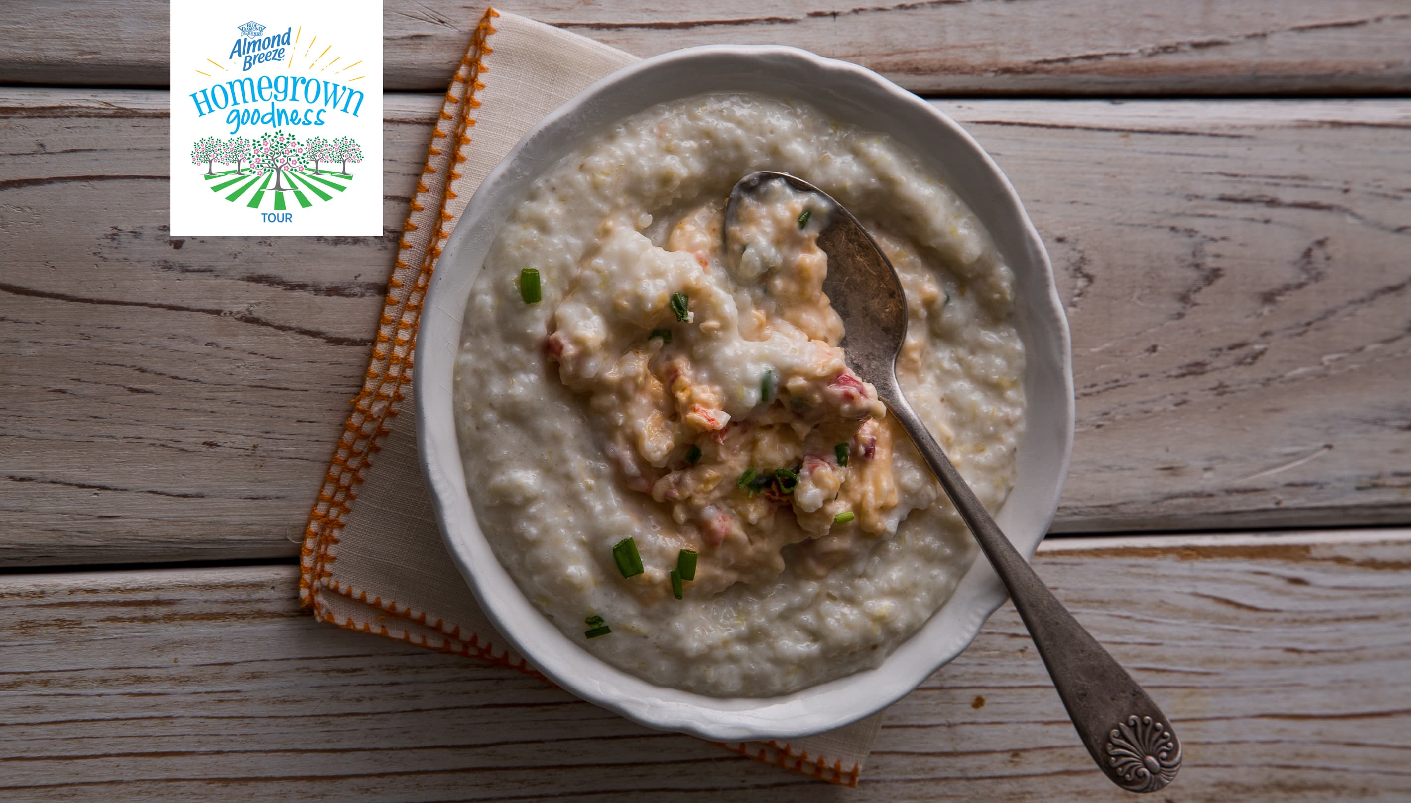 A bowl of creamy grits topped with a savory mixture and chopped herbs sits on a napkin with a spoon; a Hometown Goodness logo appears in the corner.