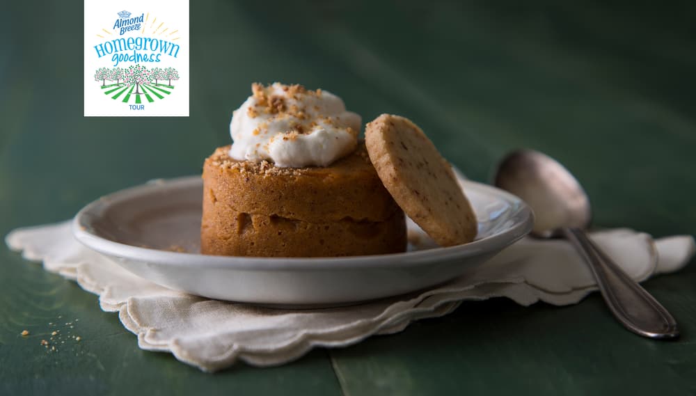 A small cake topped with whipped cream and crumbs, served with a round cookie on a white plate, next to a spoon on a cloth napkin. Hometown goodness logo in the top left corner.