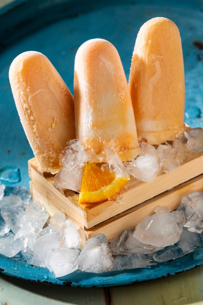 Three orange popsicles on a wooden stand with ice cubes and an orange slice, set on a blue tray.