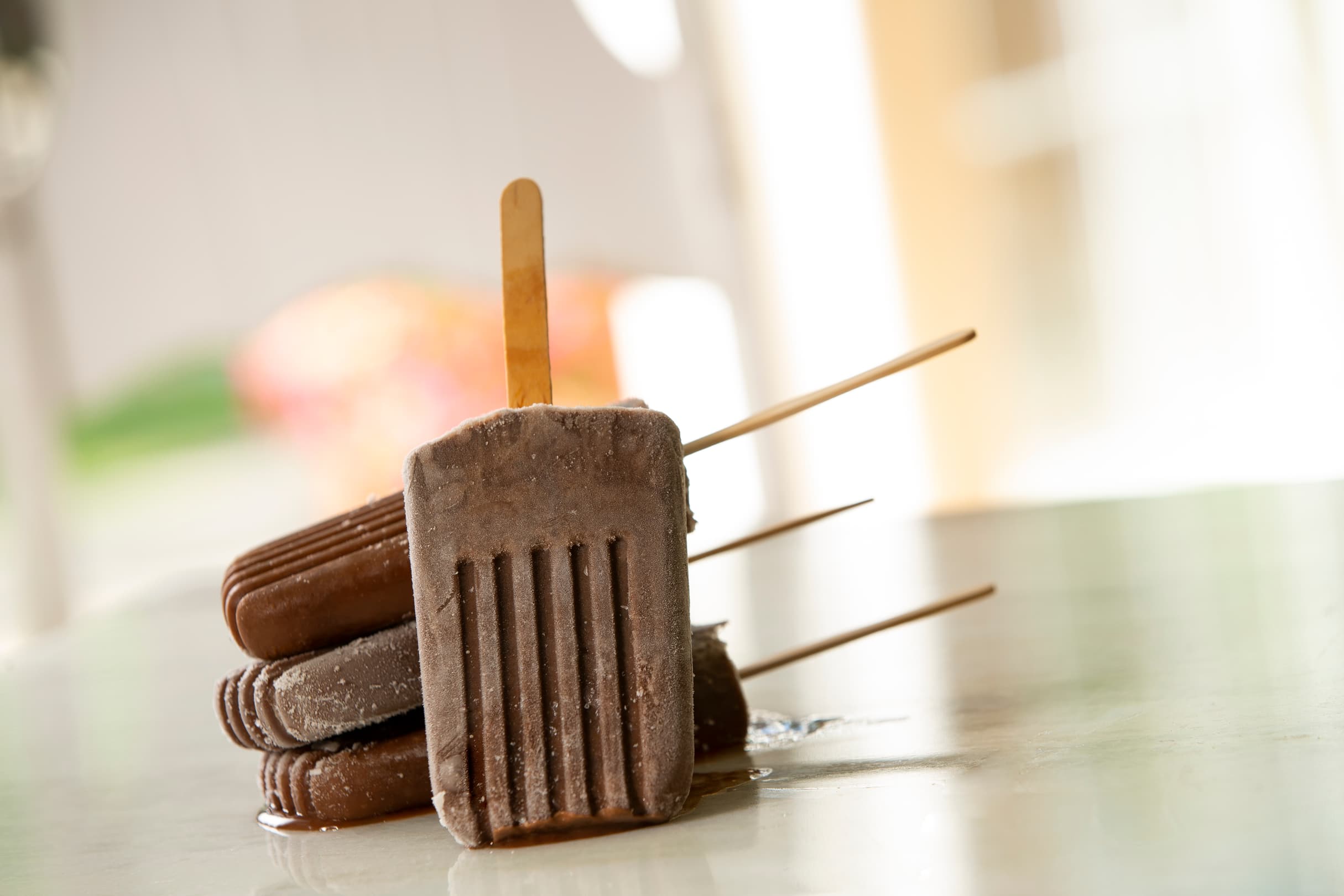 Four chocolate popsicles stacked on a shiny surface, starting to melt, with wooden sticks facing in different directions.