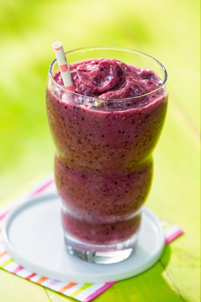 A tall glass filled with a thick, purple fruit smoothie and a paper straw, placed on a small plate with a striped napkin underneath.