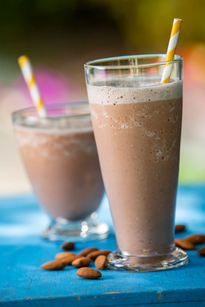 Two glasses of chocolate almond milkshake with striped straws on a blue surface, surrounded by whole almonds.