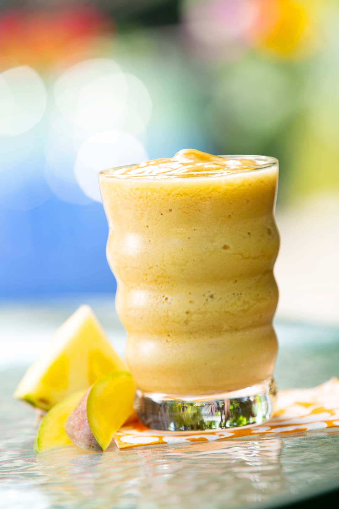A glass of yellow smoothie sits on a table next to pineapple and mango slices, with a blurred colorful background.