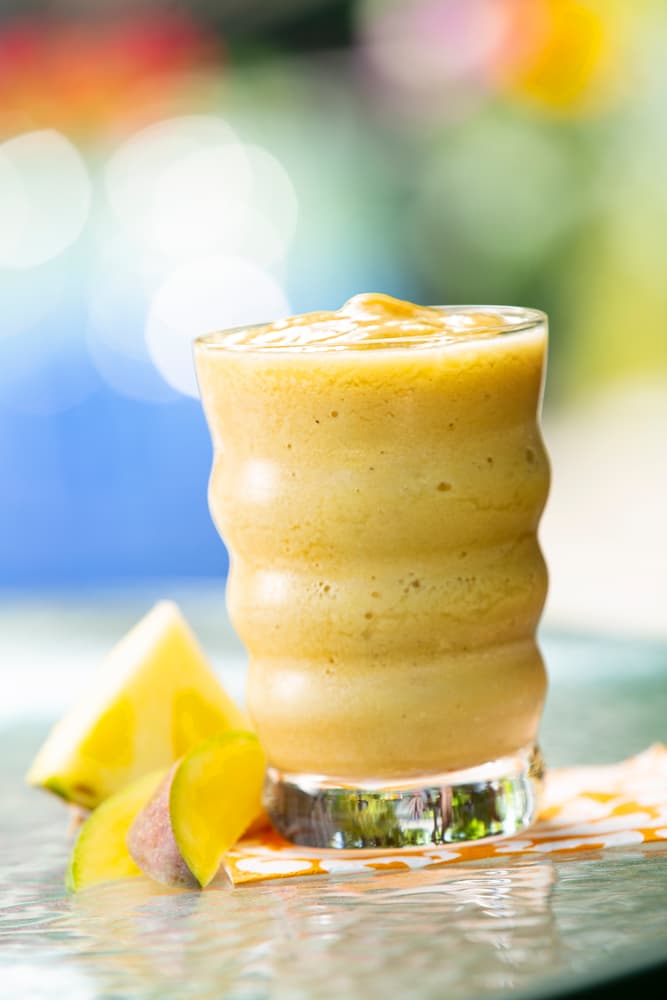 A glass of yellow smoothie sits on a table next to pineapple and mango slices, with a blurred colorful background.