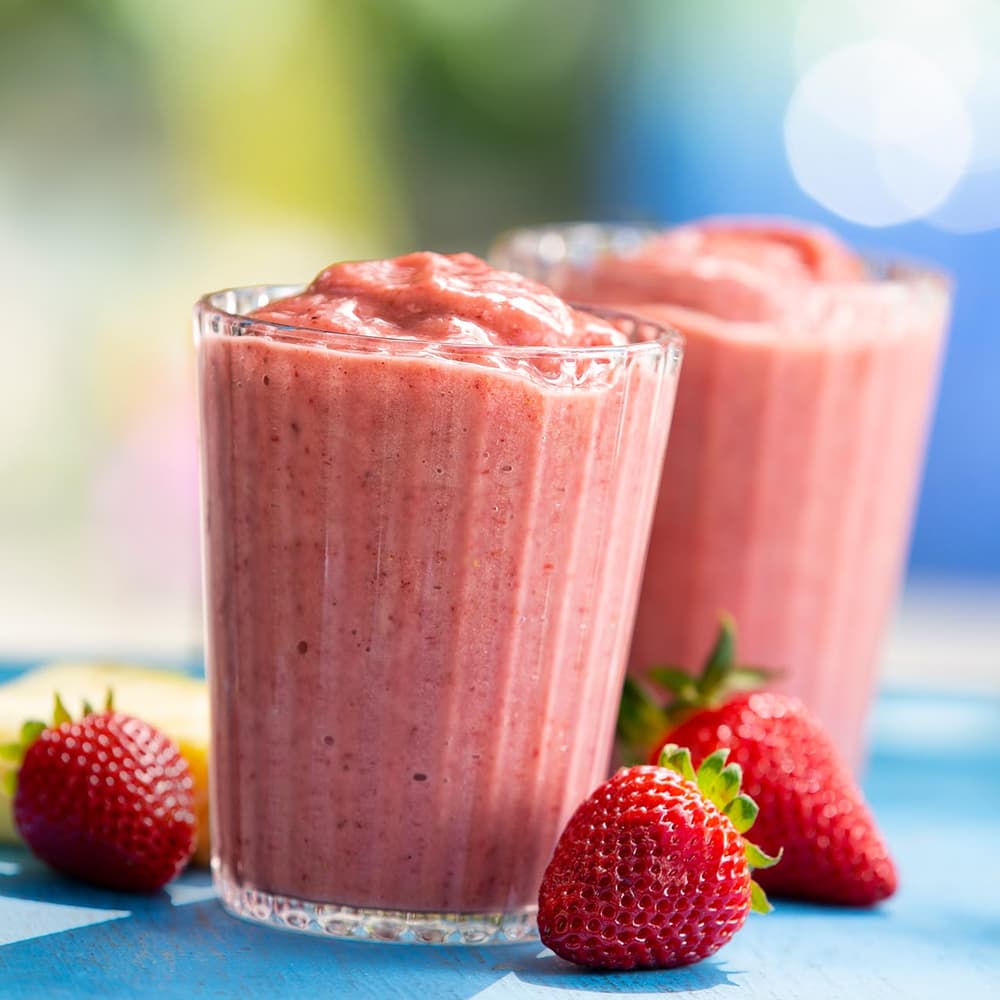 Two glasses of thick strawberry smoothie are placed on a blue surface, surrounded by whole strawberries and blurred background scenery.