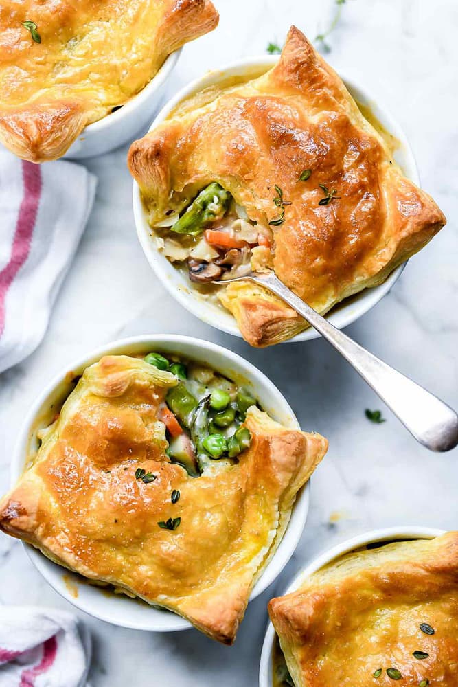 Three individual pot pies with golden-brown puff pastry crusts, filled with vegetables, on a white surface with a spoon in one of the pies.