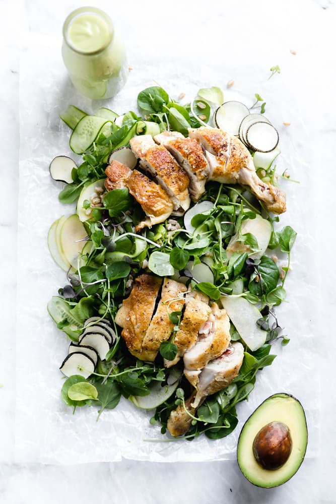 A salad with sliced roast chicken, avocado, cucumber, greens, and radish on parchment paper, with a bottle of green dressing nearby.