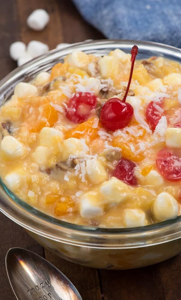 A glass bowl filled with ambrosia salad containing marshmallows, fruit pieces, shredded coconut, and topped with a maraschino cherry.