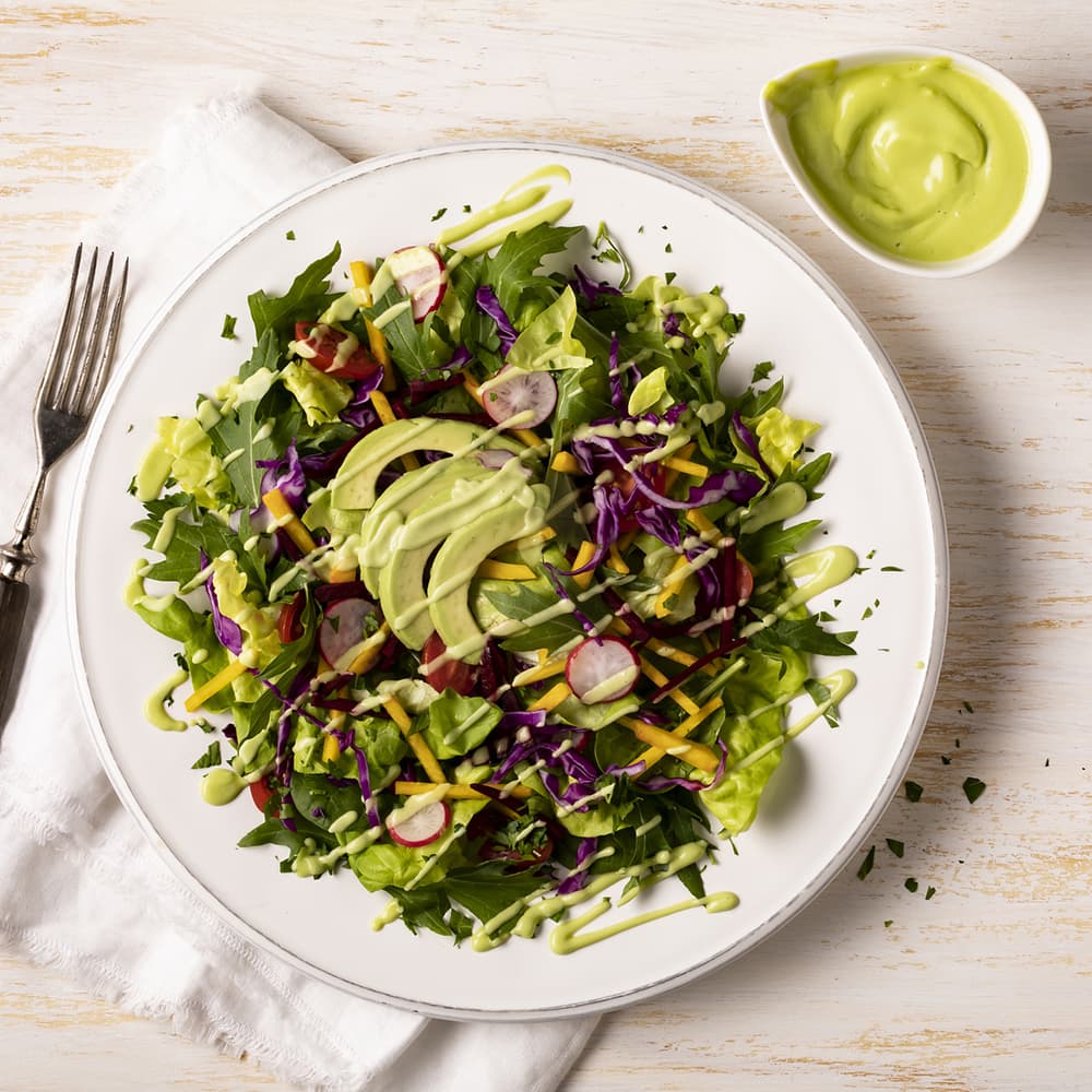 A plate of mixed green salad with avocado slices, shredded vegetables, and creamy green dressing, placed on a white napkin with a fork and a bowl of extra dressing on the side.