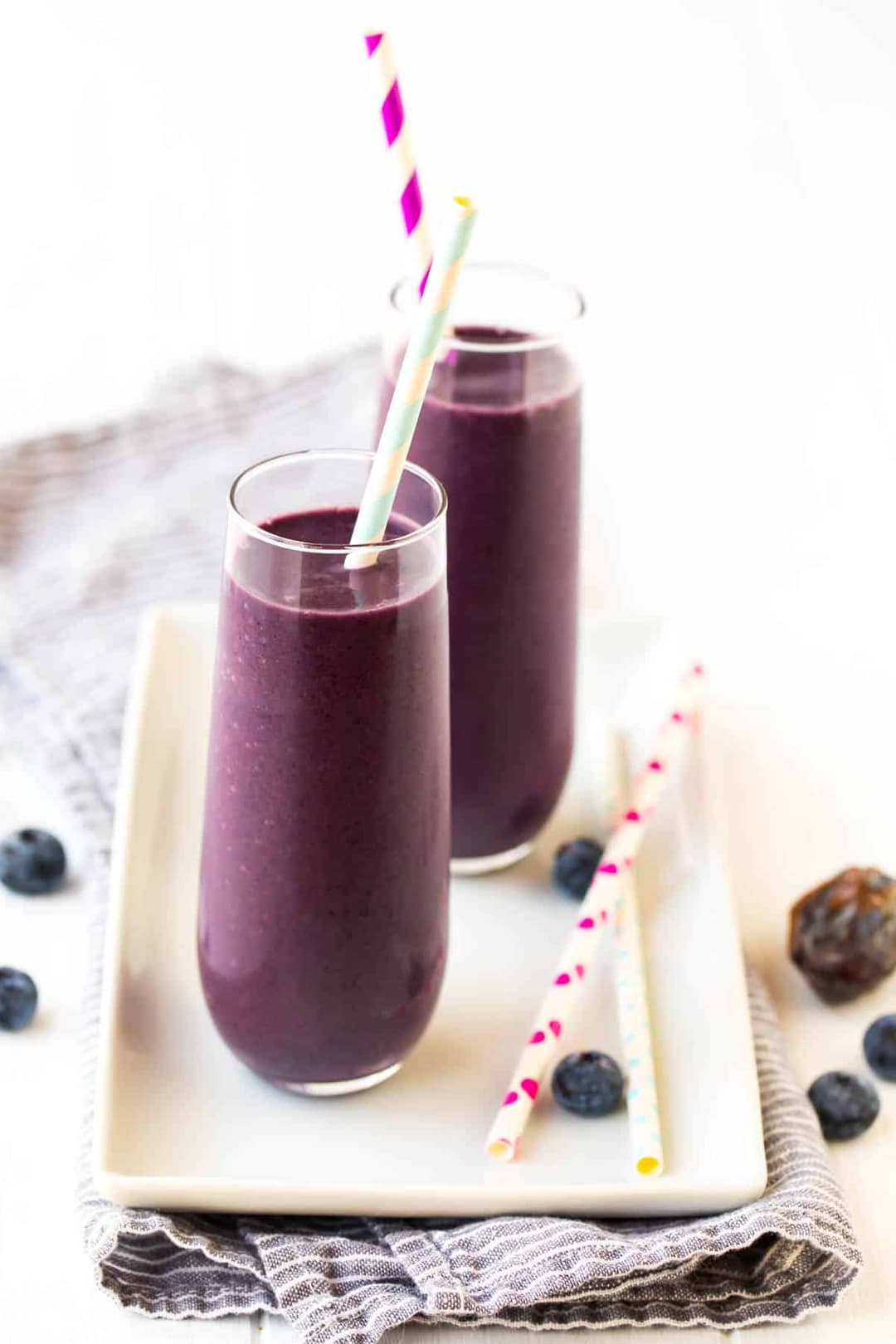Two tall glasses of purple smoothie with striped straws on a white plate, surrounded by a few blueberries and extra straws on a gray cloth.