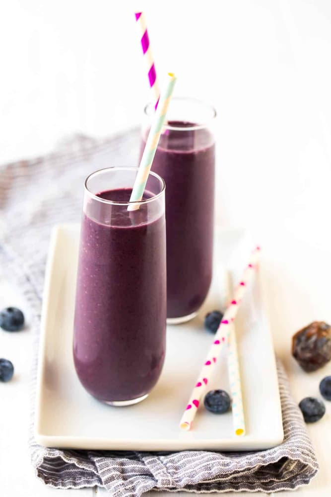 Two tall glasses of purple smoothie with striped straws on a white plate, surrounded by a few blueberries and extra straws on a gray cloth.