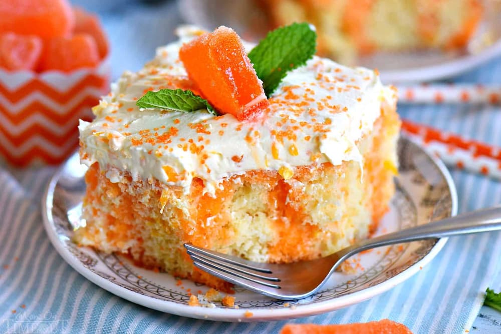 A slice of orange poke cake with white frosting, orange sprinkles, a gummy candy, and mint garnish on a plate with a fork.