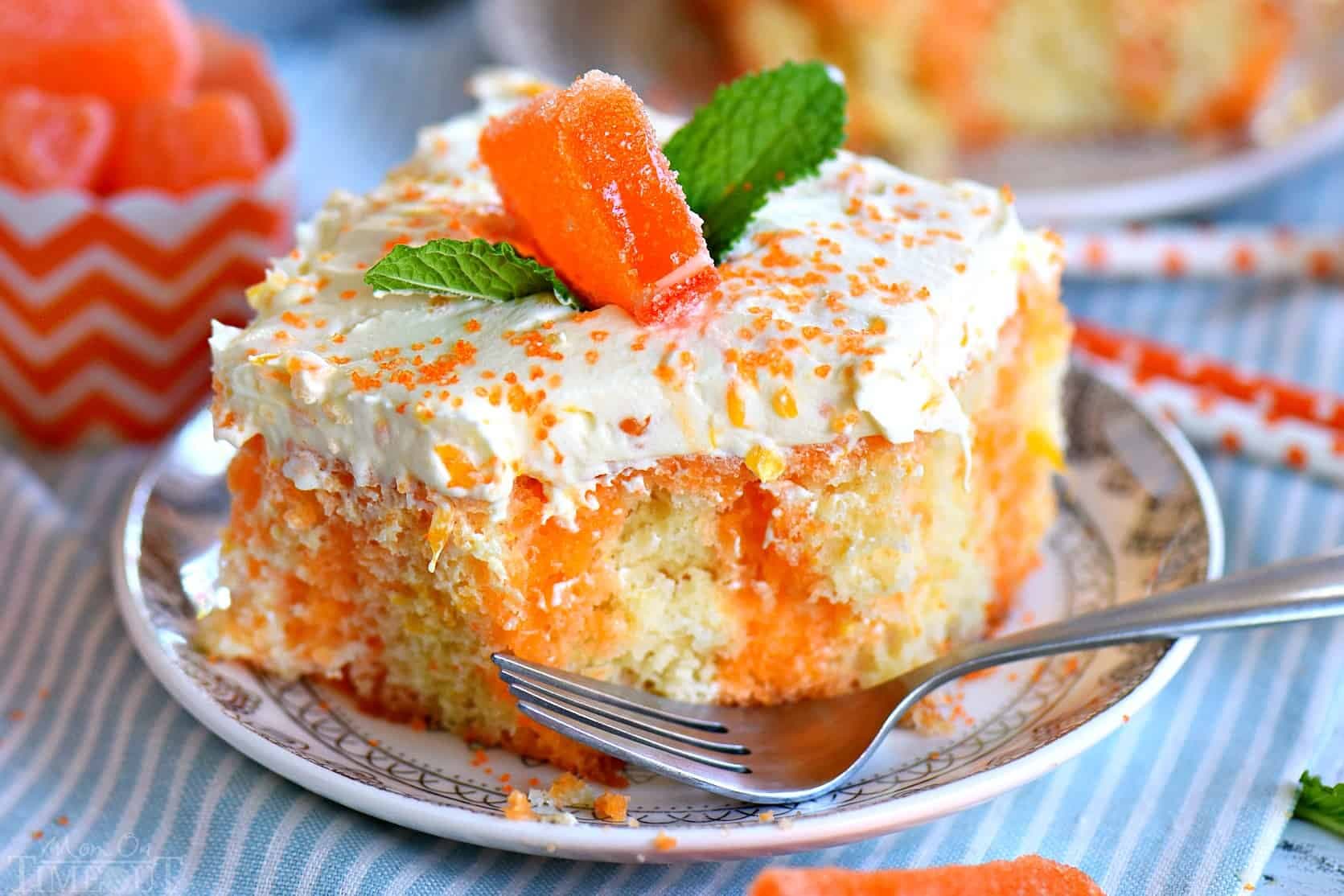 A slice of orange poke cake with white frosting, orange sprinkles, a gummy candy, and mint garnish on a plate with a fork.