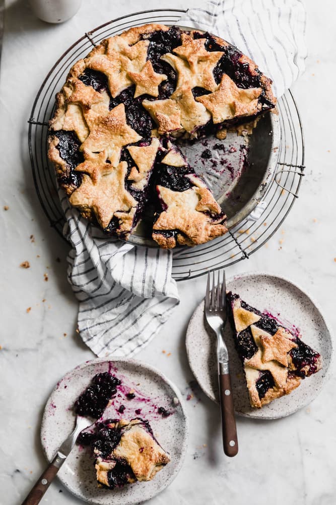 A baked berry pie with star-shaped crust on a cooling rack, with two slices served on plates, each with a fork.