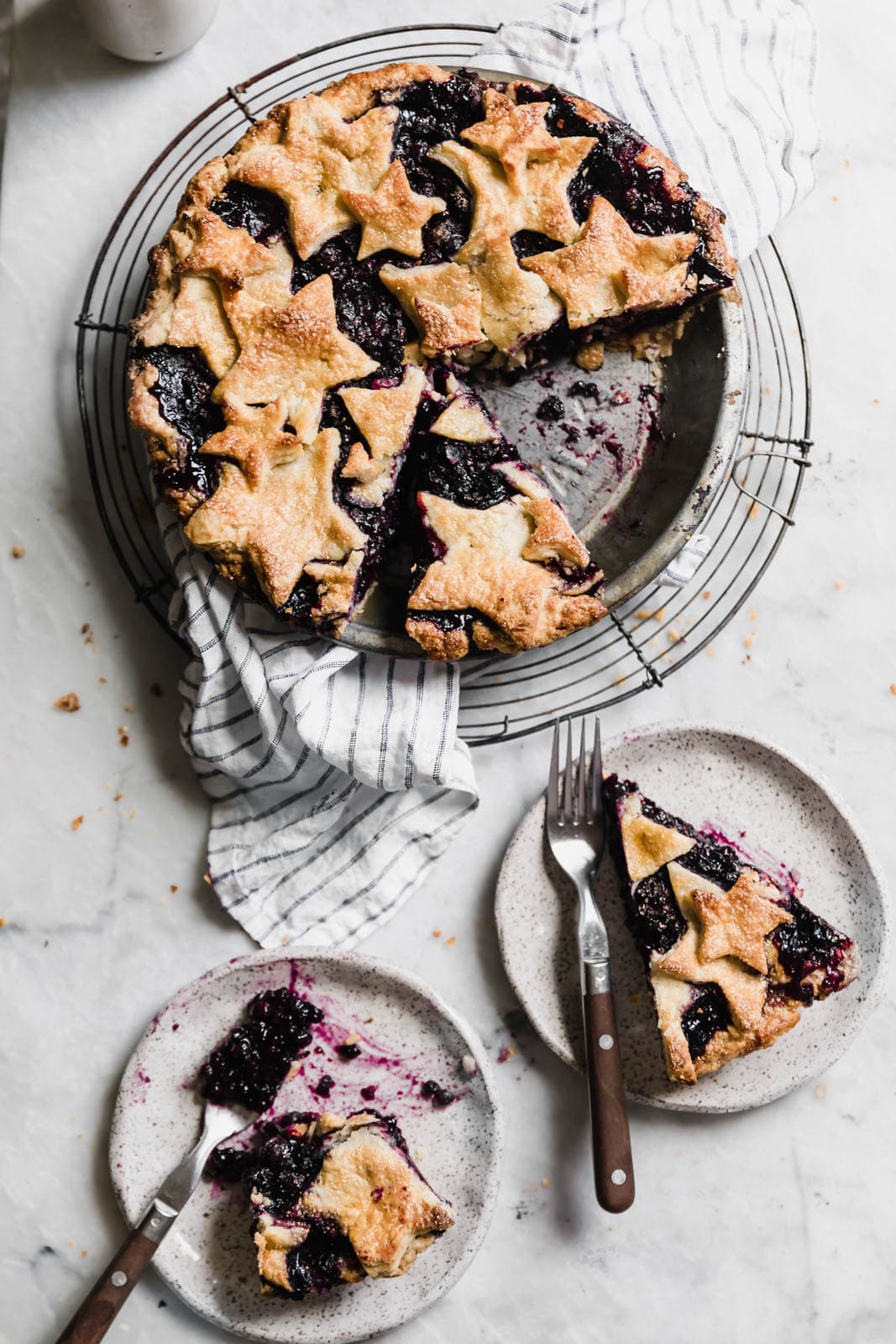 A baked berry pie with star-shaped crust on a cooling rack, with two slices served on plates, each with a fork.