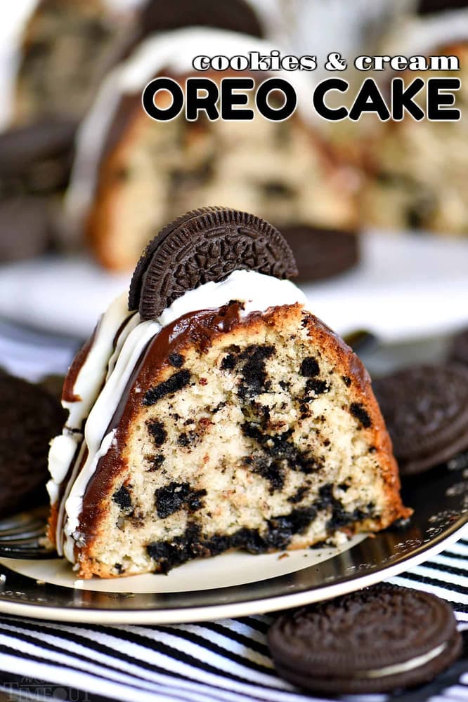 A slice of Oreo cake on a plate, topped with white icing, chocolate glaze, and a whole Oreo cookie, with more Oreos in the background.
