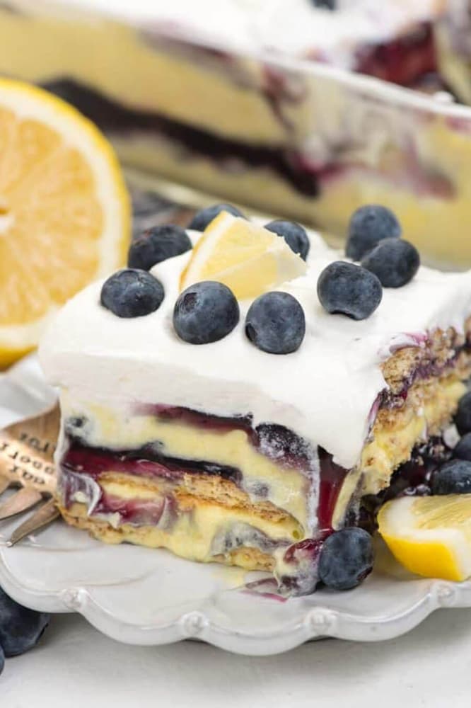 A slice of layered dessert with blueberries, lemon wedge, and whipped topping sits on a plate, with more dessert and fresh lemons in the background.