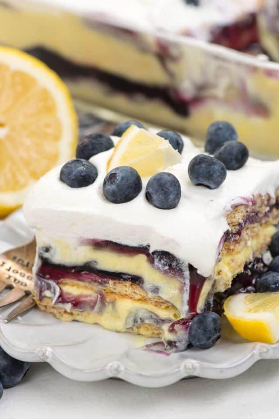 A slice of layered dessert with blueberries, lemon wedge, and whipped topping sits on a plate, with more dessert and fresh lemons in the background.