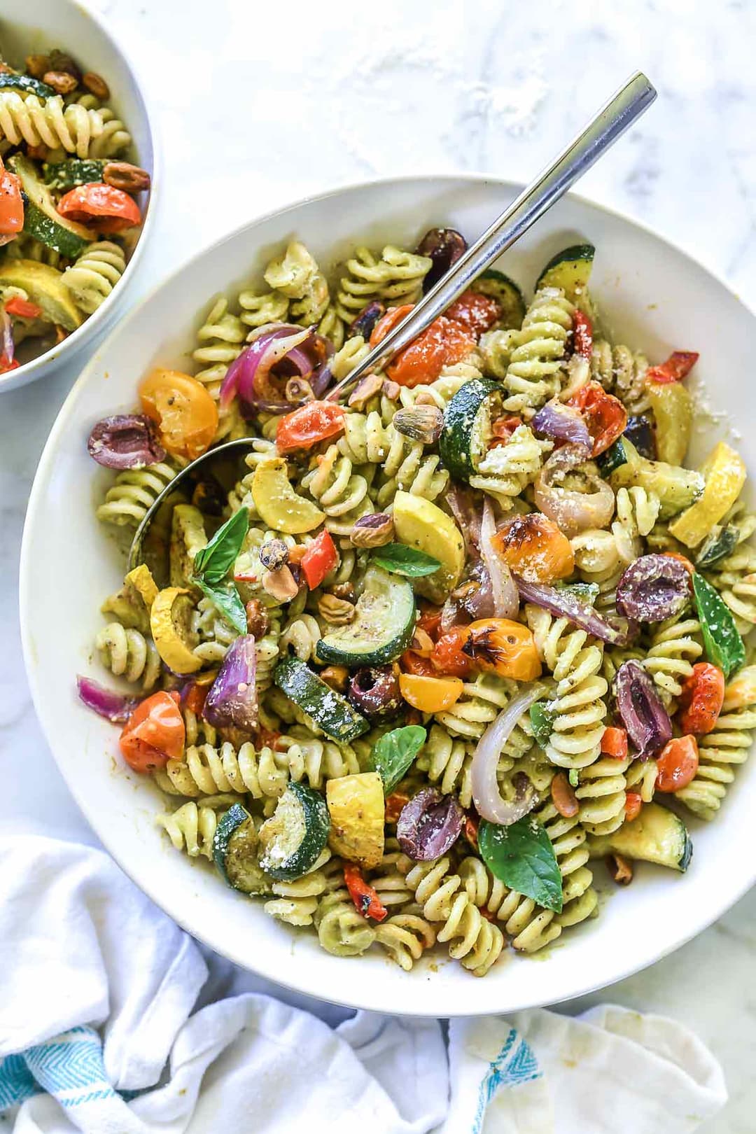 A bowl of rotini pasta mixed with roasted vegetables, including zucchini, tomatoes, red onions, and olives, garnished with fresh basil.