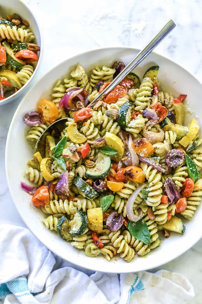 A bowl of rotini pasta mixed with roasted vegetables, including zucchini, tomatoes, red onions, and olives, garnished with fresh basil.