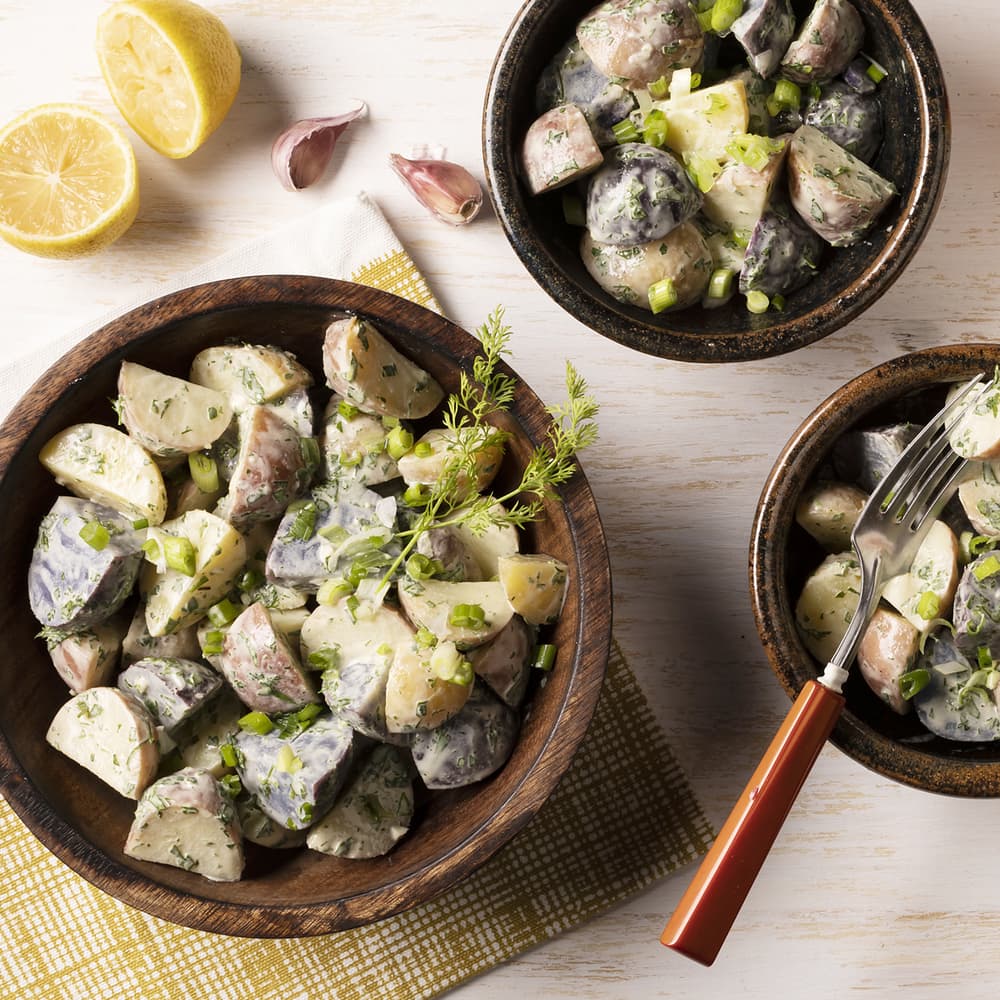 Three bowls of creamy potato salad with chopped herbs and green onions, placed on a wooden table beside halved lemons and garlic cloves. A fork rests in one bowl.