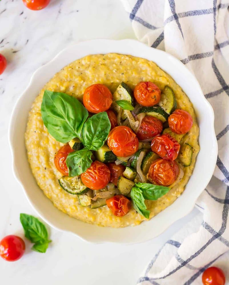 A white bowl of creamy polenta topped with roasted cherry tomatoes, zucchini, red onions, and fresh basil leaves, set on a marble surface with a checkered cloth.