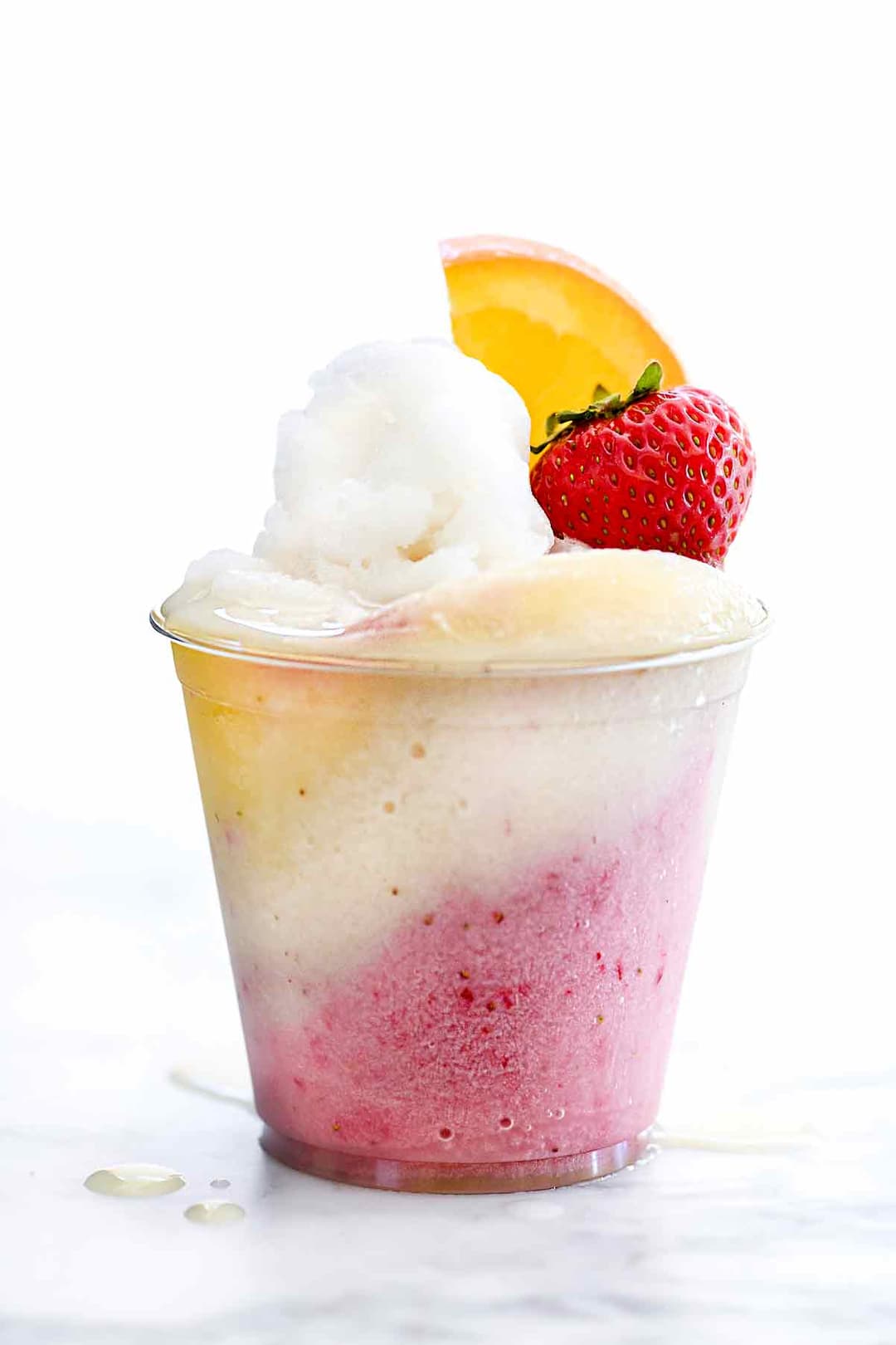 A clear plastic cup filled with pink, white, and yellow shaved ice, topped with a scoop of white ice, a fresh strawberry, and an orange slice, on a white surface.