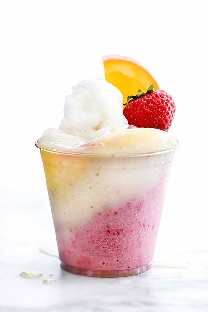 A clear plastic cup filled with pink, white, and yellow shaved ice, topped with a scoop of white ice, a fresh strawberry, and an orange slice, on a white surface.