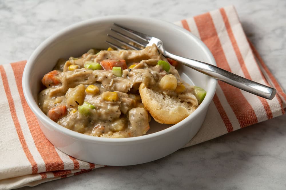 A white bowl of creamy chicken and vegetable stew served over a split biscuit, with a fork resting on the rim, on a striped napkin.