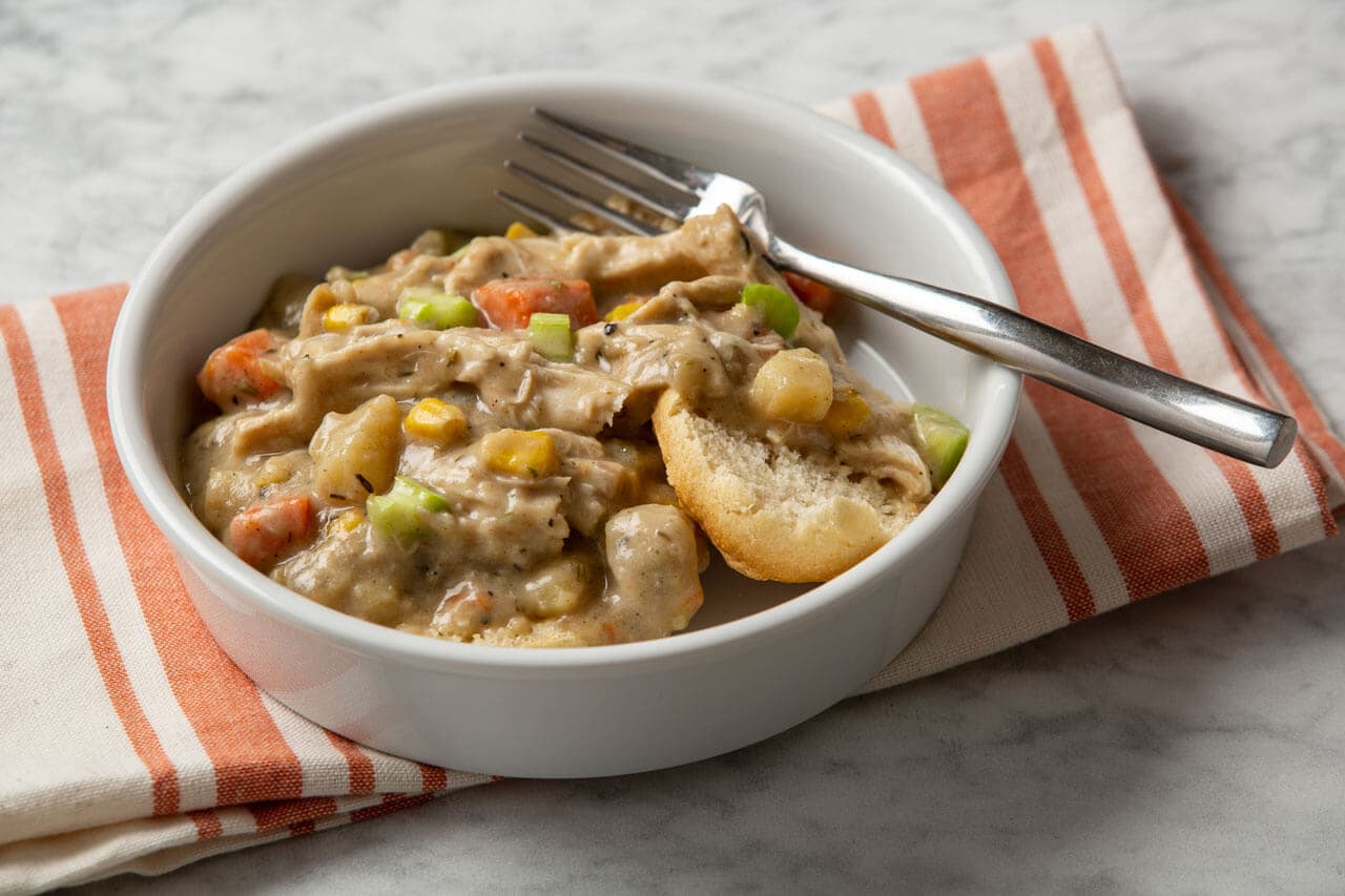 A white bowl of creamy chicken and vegetable stew served over a split biscuit, with a fork resting on the rim, on a striped napkin.