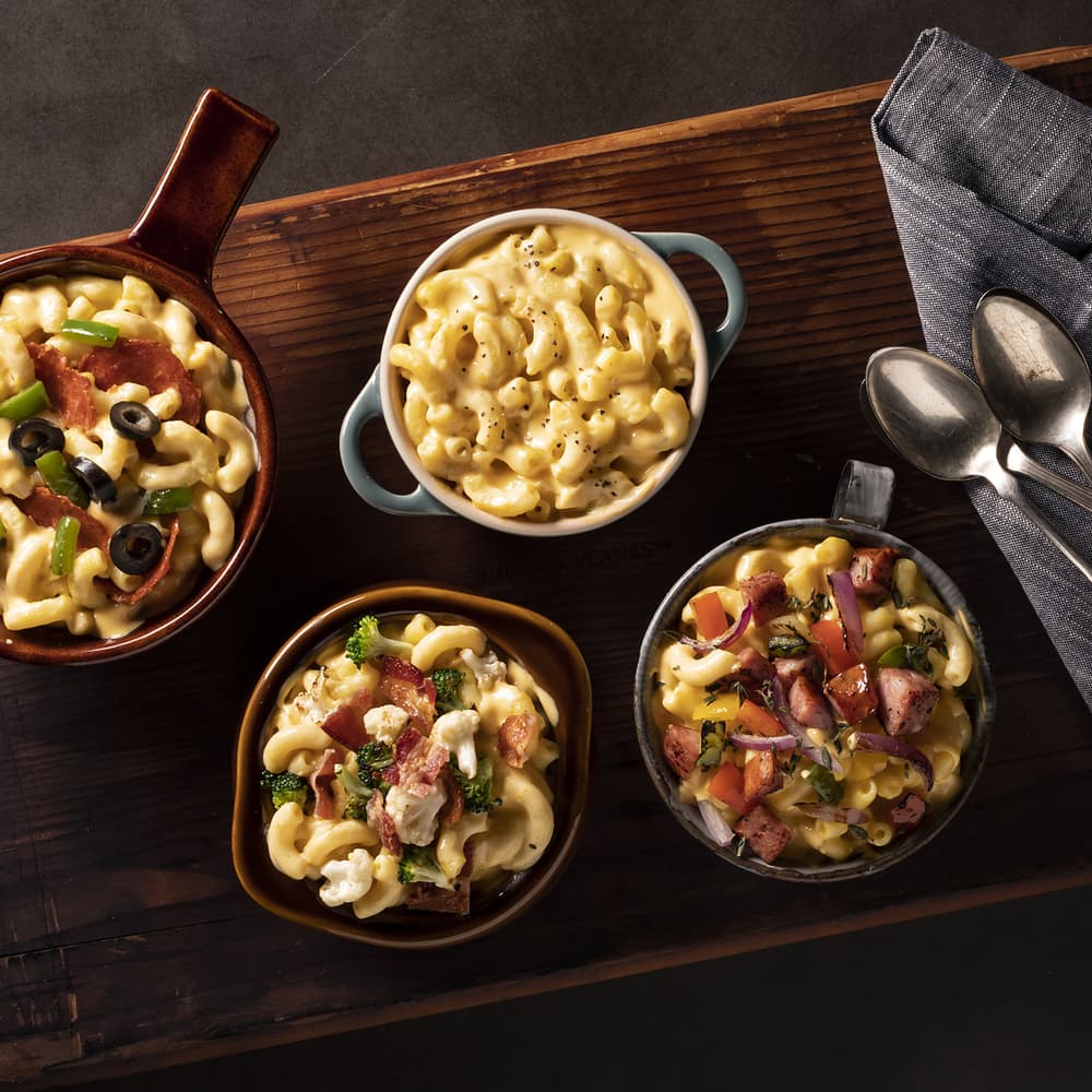Four bowls of macaroni and cheese with various toppings including vegetables, bacon, and olives, placed on a wooden board with a napkin and two spoons.
