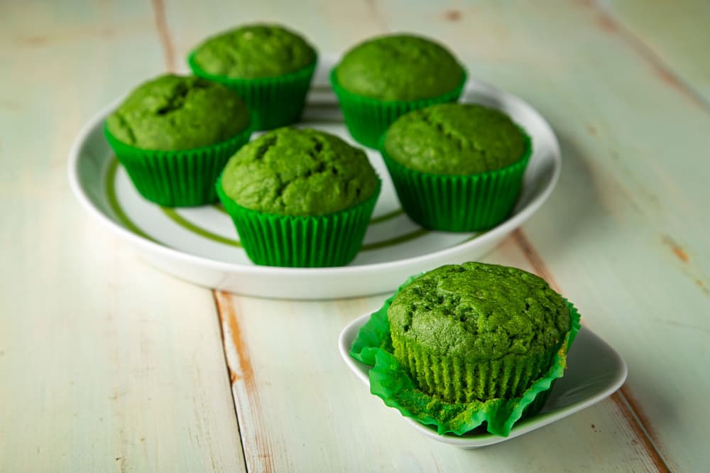 Six green muffins in green paper liners are displayed, with five on a round white plate and one on a small white dish, on a light wooden surface.