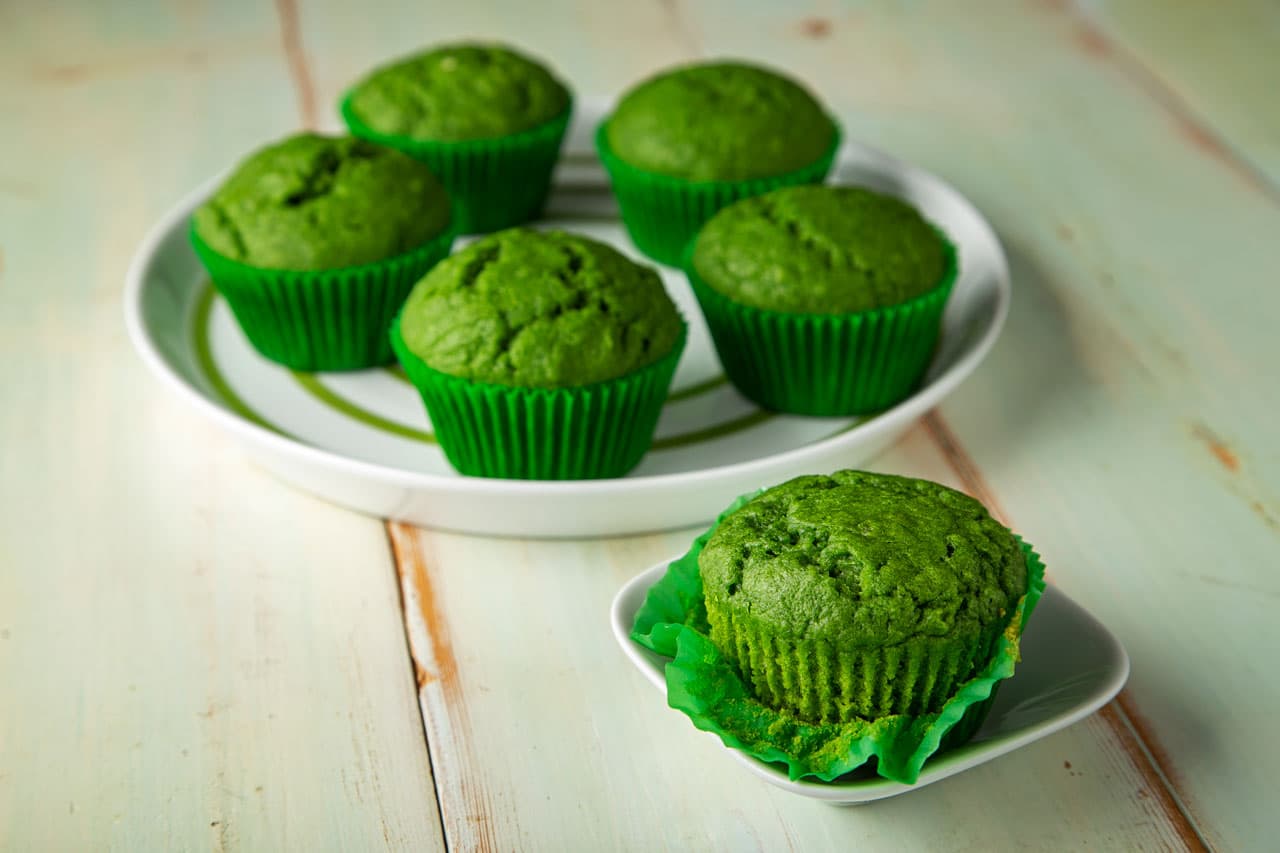 Six green muffins in green paper liners are displayed, with five on a round white plate and one on a small white dish, on a light wooden surface.