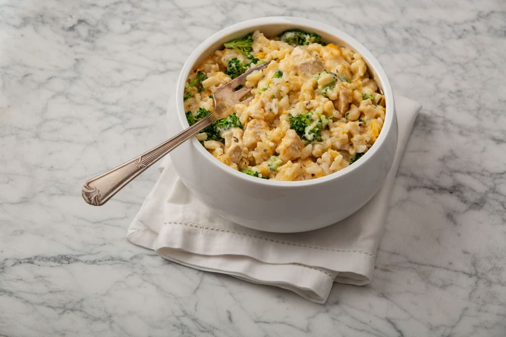 A white bowl filled with cheesy broccoli and rice casserole sits on a folded white napkin on a marble surface.