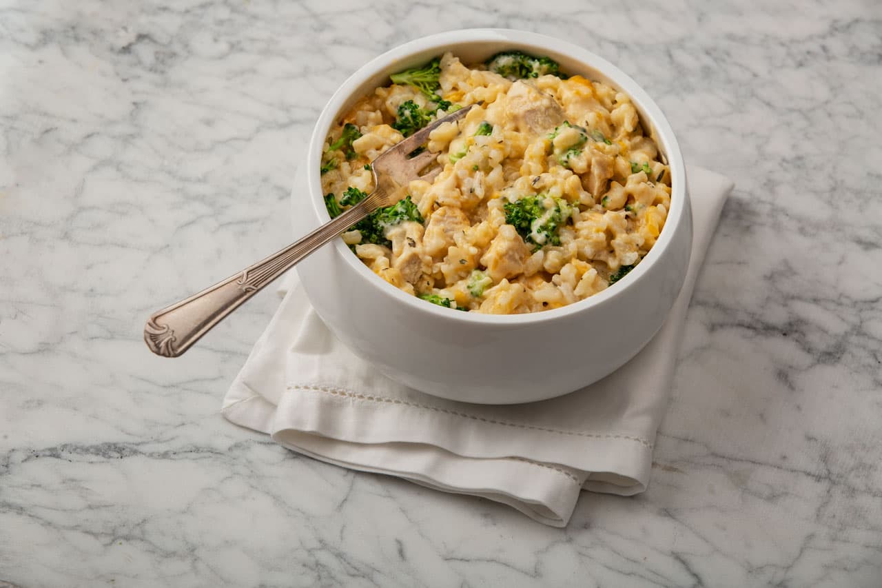 A white bowl filled with cheesy broccoli and rice casserole sits on a folded white napkin on a marble surface.
