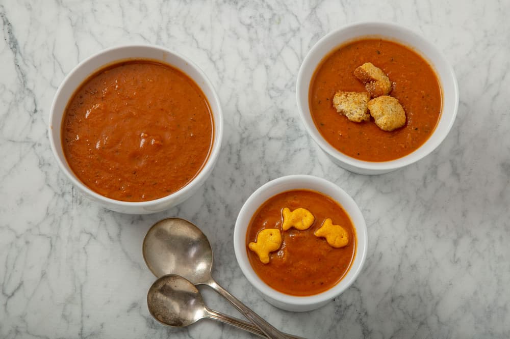 Three bowls of tomato soup on a marble surface; one with croutons, one with goldfish crackers, and three metal spoons beside them.