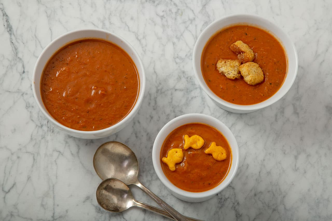 Three bowls of tomato soup on a marble surface; one with croutons, one with goldfish crackers, and three metal spoons beside them.