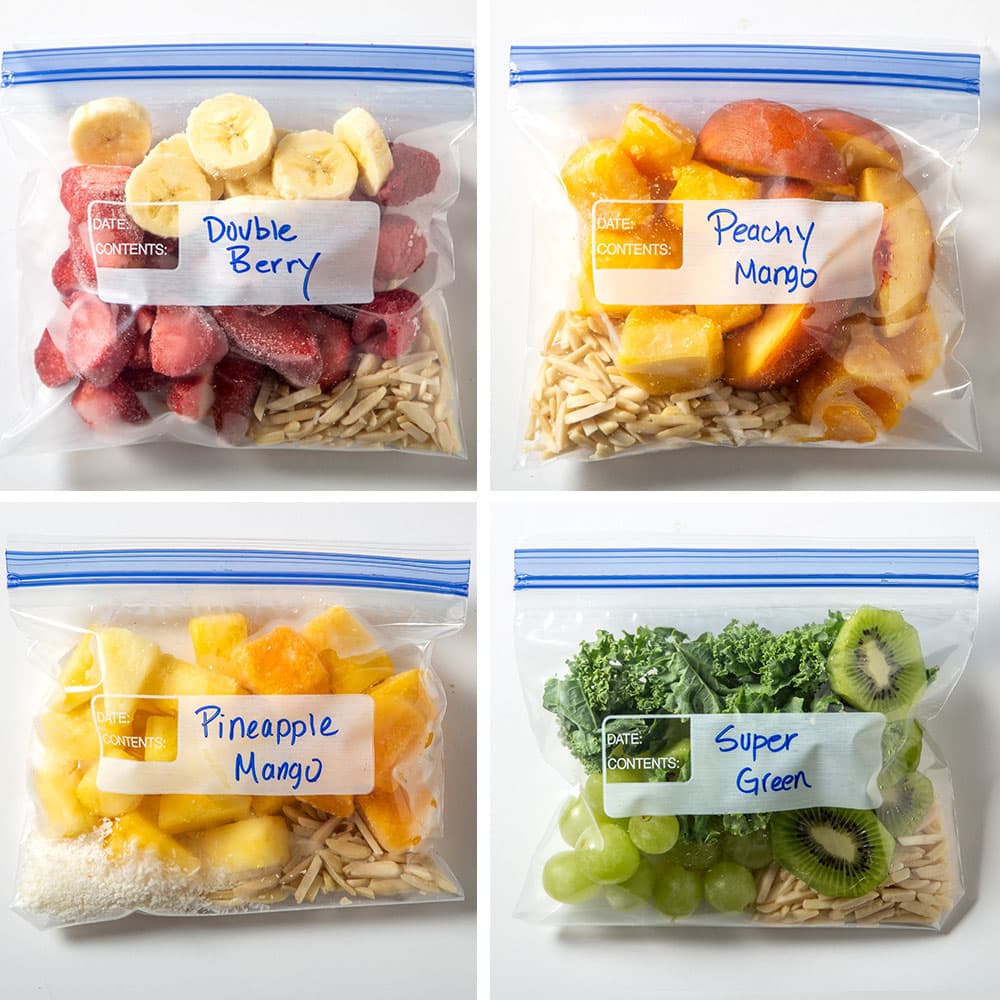 Four labeled freezer bags with fruit and veggies: Double Berry, Peachy Mango, Pineapple Mango, and Super Green, each containing assorted sliced produce ready for smoothies.