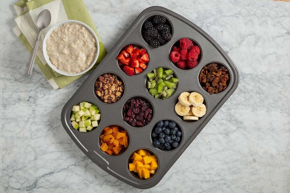 A muffin tin holds various fruits, nuts, and toppings on a marble surface next to a bowl of oatmeal and a spoon on a folded napkin.