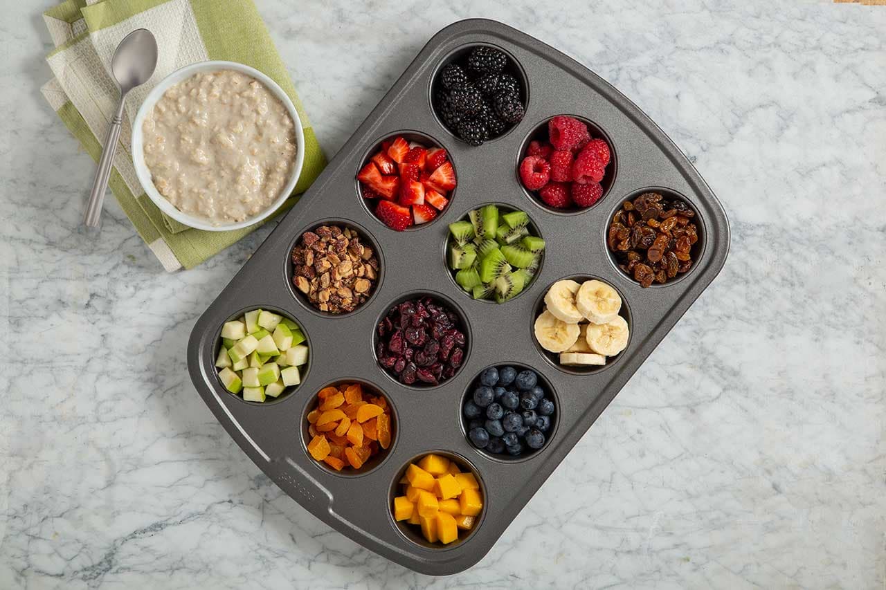 A muffin tin holds various fruits, nuts, and toppings on a marble surface next to a bowl of oatmeal and a spoon on a folded napkin.