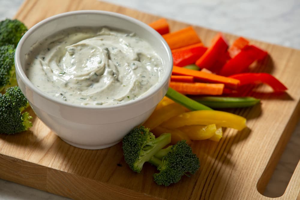 A bowl of creamy dip sits on a wooden board next to sliced carrots, red and yellow bell peppers, green beans, and broccoli florets.
