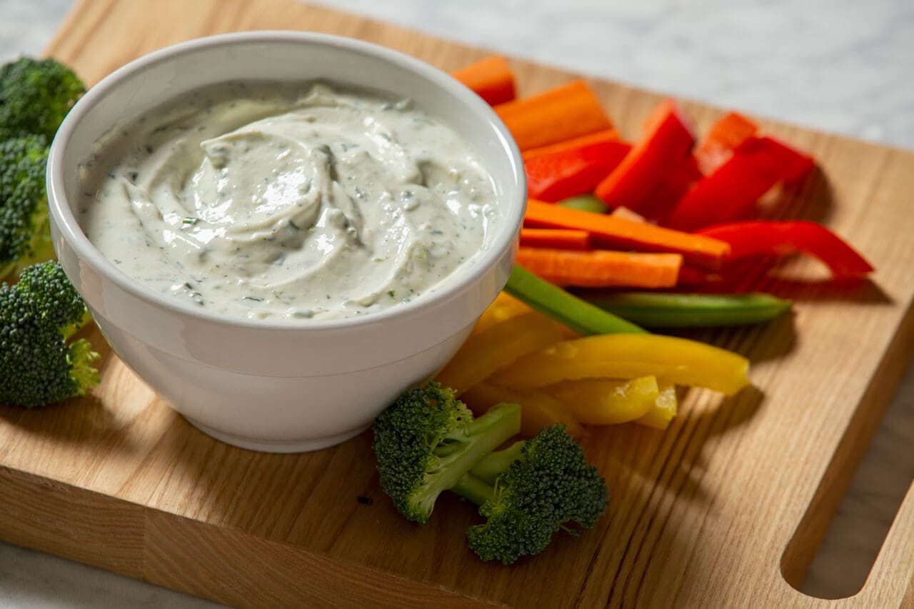 A bowl of creamy dip sits on a wooden board next to sliced carrots, red and yellow bell peppers, green beans, and broccoli florets.