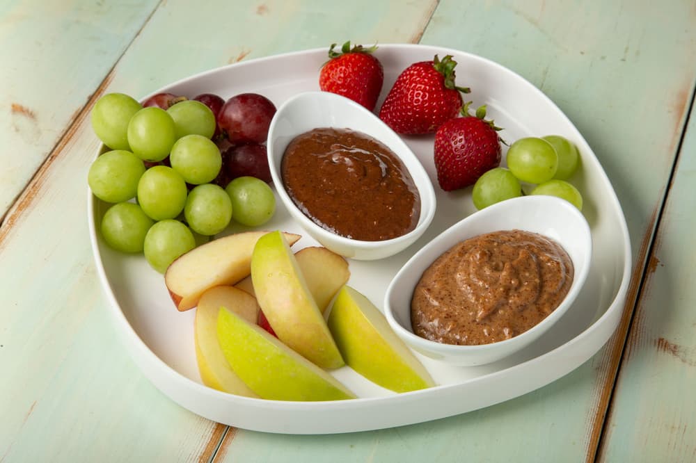 White plate with green grapes, red grapes, apple slices, strawberries, and two small bowls of brown dips on a light wooden table.